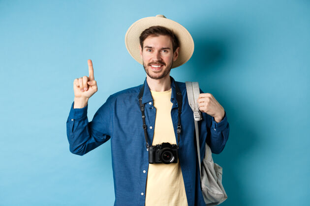 帅气帅气的快乐旅游者 戴着夏帽 背着背包 拿着相机 手指着logo 推荐旅行社或度假地 蓝色背景年轻肖像成人