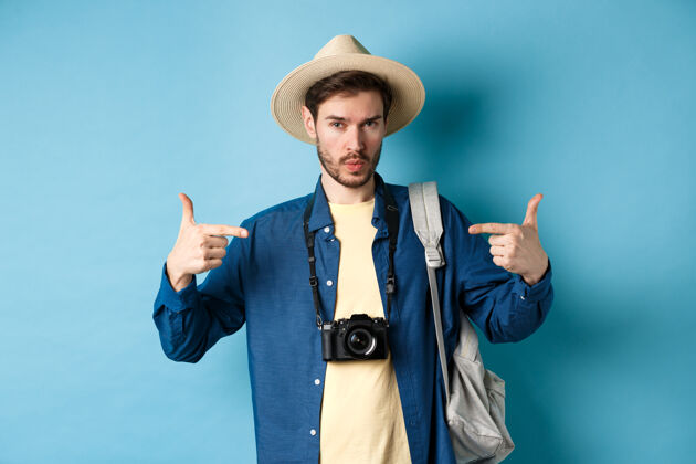 人自信而有准备的年轻人指着自己 自我推销或炫耀 去放暑假 戴草帽 背着带相机的背包 蓝色背景男男朋友
