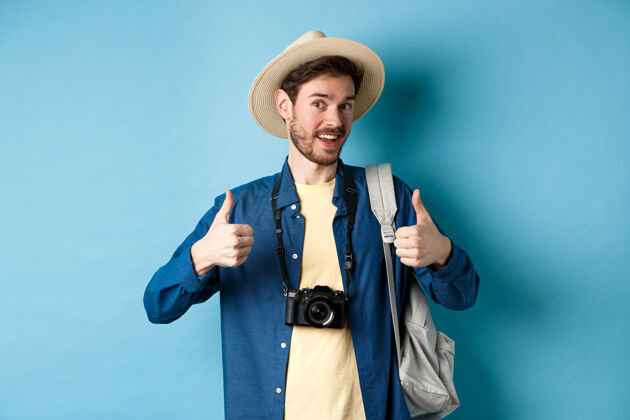 认可快乐微笑的游客说是的 竖起大拇指 去放暑假 拿着背包和照相机 蓝色的背景喜欢男成人