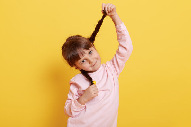 表情有趣的小女孩扎着辫子 带着可爱的微笑直视镜头 穿着淡粉色的衬衫 可爱的孩子靠在黄色的墙上乐观乐趣孩子