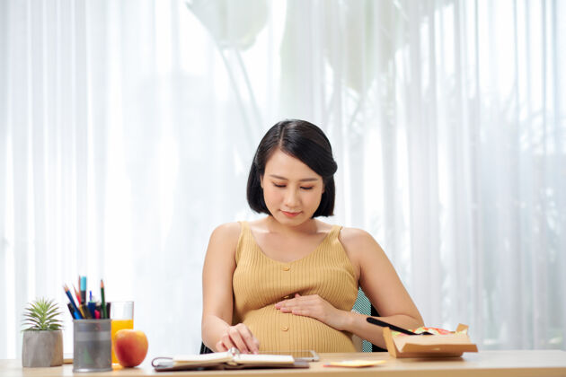 产妇怀孕的年轻女子坐在客厅的桌子上使用数码平板电脑 吃早餐和工作怀孕女人护理