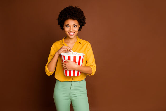 深色头发照片中迷人的深色皮肤波浪女士拿着爆米花桶吃玉米看最喜欢的电视节目穿黄色衬衫绿色裤子孤立的棕色皮肤女性Afro
