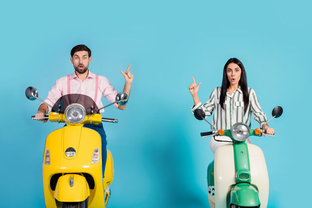 情侣惊讶的两个人妻子丈夫骑摩托车骑黄绿色摩托车印象深刻的广告看旅行点食指文案穿条纹粉色衬衫隔离蓝色墙壁手势骑手骑行