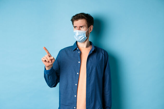 面具Covid-19和医疗保健概念困惑和怀疑的人戴着面具皱着眉头 指着空荡荡的空间 站在蓝色的背景上流行病冠状病毒男性