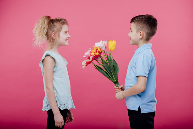 童年两个小孩小男孩把花伸向一个穿着粉色连衣裙的小女孩 隔离在蓝色的墙上 侧面图侧写情感风景