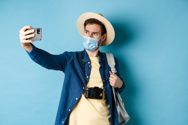 自信Covid-19 流行病和旅行概念暑假的游客戴着医用面罩自拍 用智能手机拍照 站在蓝色背景上相机肖像朋友