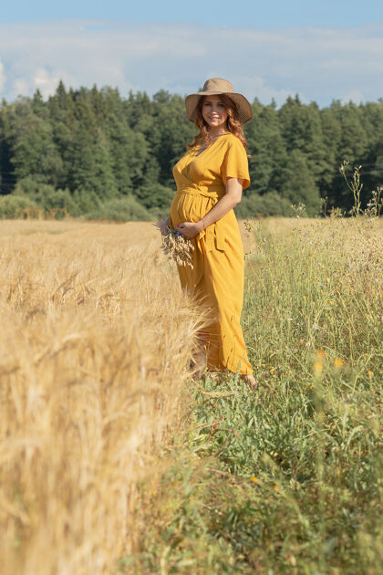 可爱在一个阳光明媚的夏日 一位年轻漂亮的孕妇穿着黄色的裙子 戴着黄色的帽子 走过一片麦田肖像举行肚子