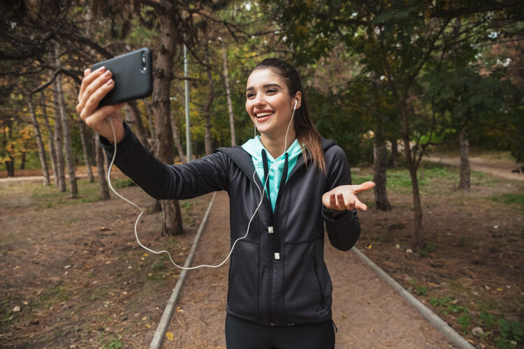 运动微笑的健身女士戴着耳机听音乐 站在公园里自拍休息深色头发运动