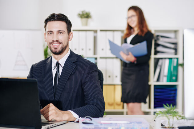 办公桌在办公桌上用笔记本电脑工作的快乐的年轻企业家的肖像 他的助手在背景中寻找文件西装团队合作女性