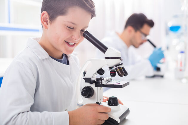 学习看一看可爱聪明的小男孩一边看着显微镜一边微笑学习者设备纯洁性