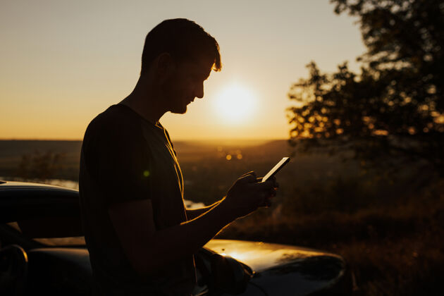 人壮丽的日落时分 坐汽车和拿着手机站在山上的男人的剪影河流华丽户外