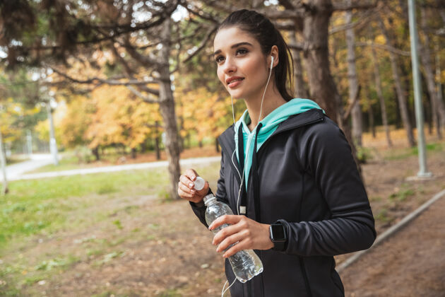 连接微笑的健身女 戴着耳机听音乐 站在公园里拿着一瓶水仅限女性运动公园
