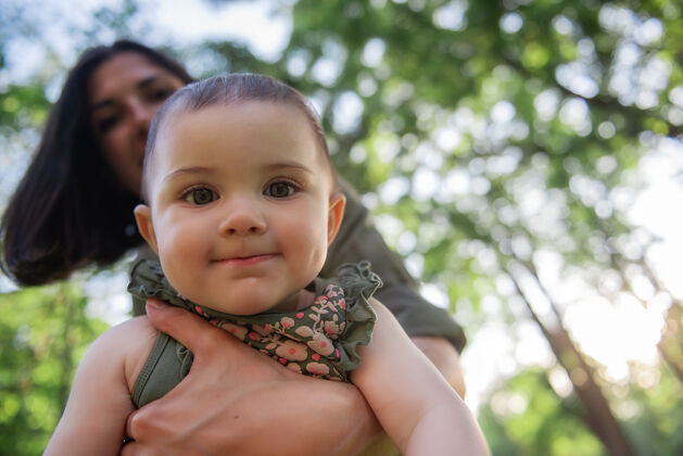 微笑年轻的棕色头发的母亲在公园里的绿树丛中摇动着怀里的孩子草女人抱着小女孩玩母亲女孩童年
