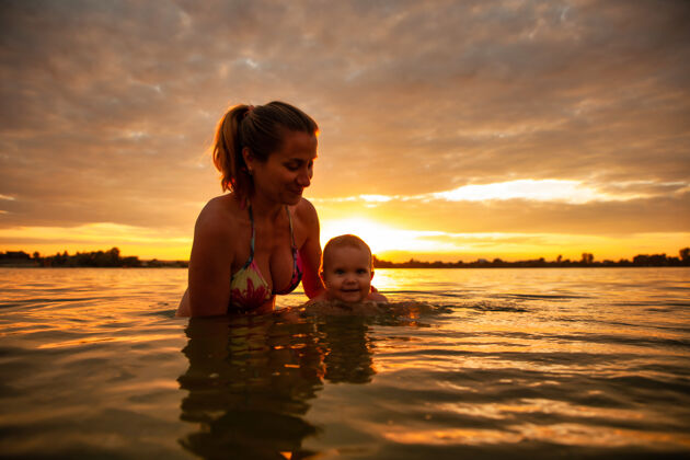 女儿快乐的白种人的母亲教游泳小可爱的微笑的婴儿在海水中的侧视图人日落水