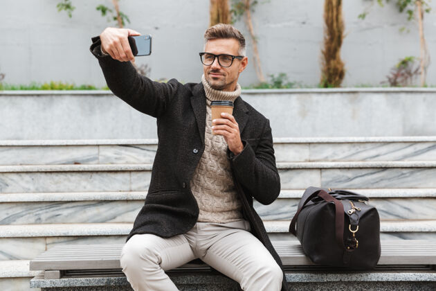 毛衣穿着夹克的帅哥坐在户外自拍 手里拿着一杯外卖咖啡休闲户外吸引