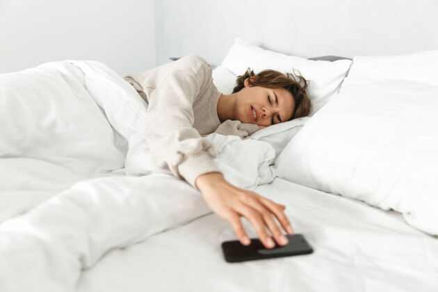 设备迷人的年轻女孩在床上放松 早上 伸手拿手机毯子吸引打字