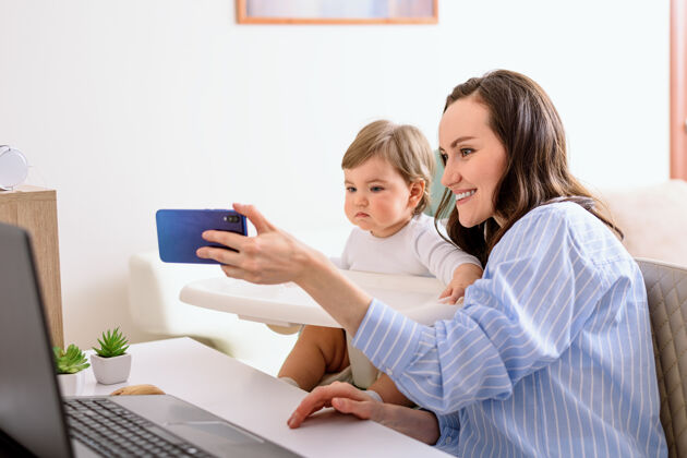 坐着穿蓝衬衫的妈妈和女儿通过视频聊天 视频通话 自拍 做妈妈笔记本电脑学习在家工作