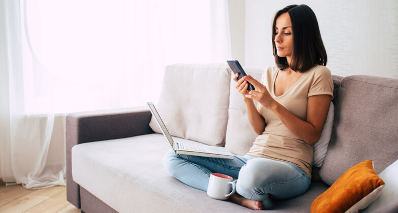工作年轻的现代自信微笑的女人在家工作或放松 坐在沙发上用智能手机休闲互联网生活