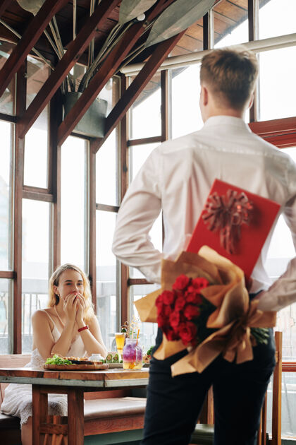 团聚兴奋的年轻女子看着男友 手里拿着一盒巧克力和鲜花来到她的桌边感受约会优雅