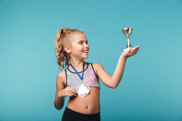 第一快乐的小体育女孩庆祝战胜蓝墙 戴着金牌 展示着奖杯冠军行动比赛