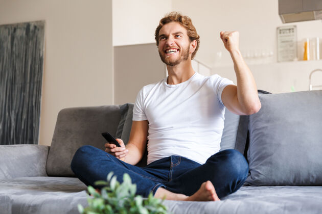 电视照片中 一位帅哥情绪激动地坐在家里的沙发上 拿着遥控器看电视 做出赢家的手势远程男人软