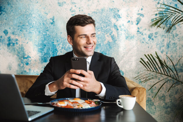 帅哥一个开朗英俊的年轻商人坐在咖啡馆里用笔记本电脑吃早餐或晚餐用手机吃饭的画面打字咖啡馆高管