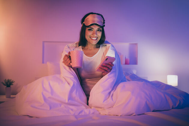 快乐可爱可爱的女人坐在床上拿着手机杯的画像杯子使用配件