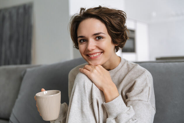 沙发快乐的年轻女子坐在家里的沙发上 手里拿着杯子放松舒适微笑