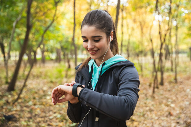 女性站在公园里 微笑着戴着耳机听音乐 查看智能手表运动员耳机公园