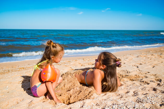 童年在一个阳光明媚温暖的夏日 快乐的小女孩在海边的沙滩上玩耍时 把姐姐埋在沙滩上休闲可爱热带