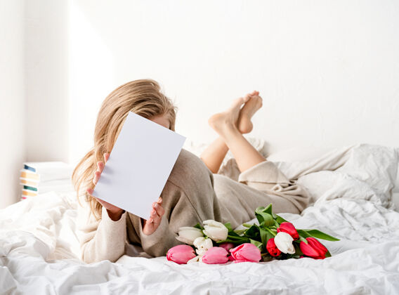 节日快乐的女人穿着睡衣躺在床上 手里拿着郁金香花束和一张设计模型的空白卡片室内脸生活方式
