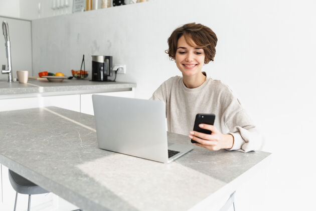 肖像微笑的年轻女子坐在厨房里 手里拿着手机 手里拿着笔记本电脑微笑电话无线