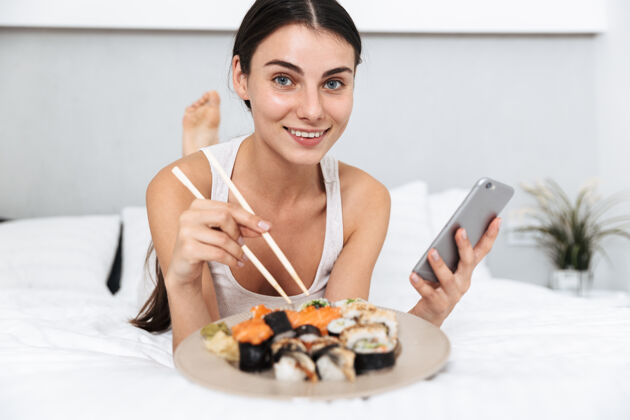 室内美丽的年轻女子在家里的床上放松 拿着手机 吃着盘子里的寿司打字床年轻