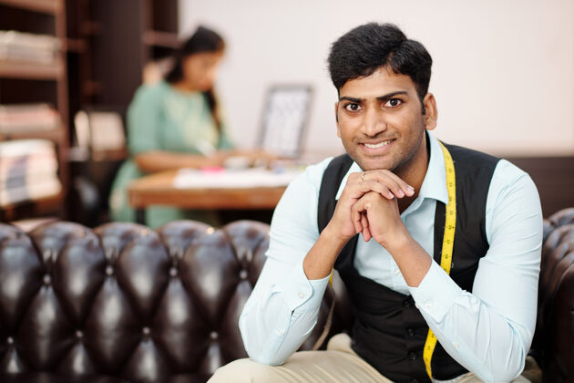 成人在工作室的沙发上 微笑的年轻印度裁缝拿着卷尺的画像裁缝商务年轻人
