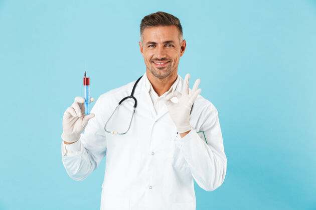 实验室自信的男医生穿着制服 孤立地站在蓝色的墙上 展示着一个装有红色液体的注射器职业成人制服