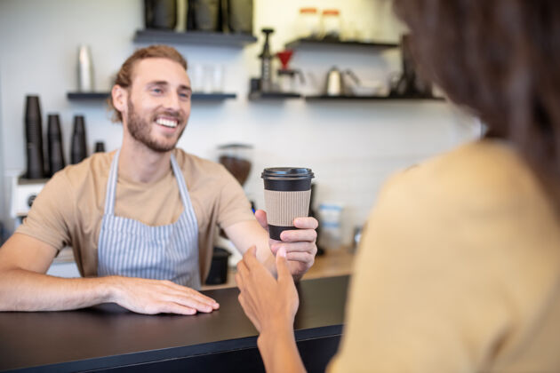 初创公司最好的咖啡年轻一个穿着围裙 背对着镜头 拿着一杯咖啡的成年男子站在吧台上对女人微笑着顾客自营职业金发