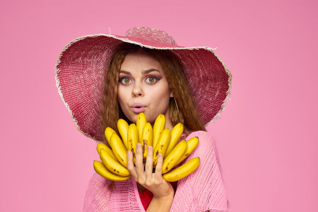 饮料女人手里拿着香蕉 戴着奇异水果的帽子 生活在粉色的墙上身体季节姿势