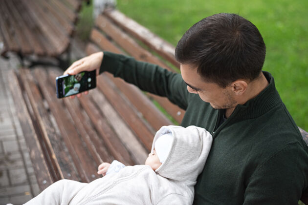 肖像穿着绿色套头衫的父亲抱着孩子坐在长凳上用智能手机自拍移动散步快乐