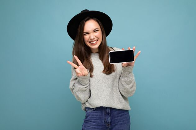 设备相当积极的年轻女性 戴着黑色帽子 穿着灰色毛衣 手持手机 背景上显示智能手机 显示和平姿态微笑使用学生