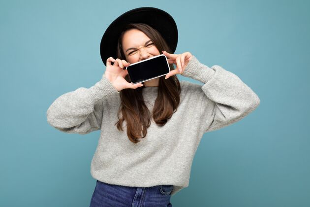 手机照片中 戴着黑色帽子 穿着灰色毛衣的美女手持手机 背景上显示智能手机被隔离在一起眼睛模仿向上 切断 自由空间手机毛衣黑帽子