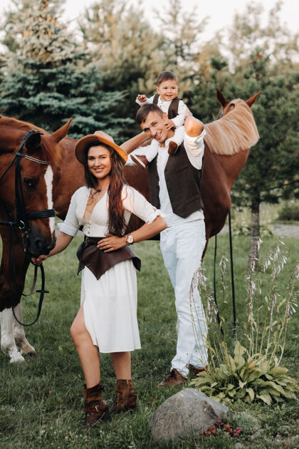 童年一个穿着白色衣服的家庭和他们的儿子站在大自然中两匹美丽的马旁边一对时髦的夫妇带着一个孩子与马合影马术夫妇与儿童女人