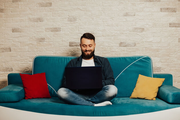舒适满脸胡须的笑脸男子一边看着笔记本电脑屏幕 一边坐在家里的绿松石沙发上专注学生工作
