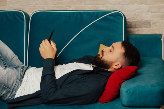 工作场所一个留着大胡子的年轻人在家上网 躺在沙发上用手机看新闻工作学习专注