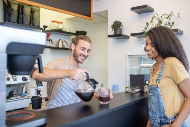 腰向上欢迎来到咖啡仁慈的微笑的男人把咖啡倒进柜台后面的杯子里 等待着快乐的混血女女人忙碌杯子