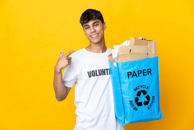 年轻一个男人拿着一个装满废纸的回收袋 翻过隔离的黄色墙壁 竖起大拇指好手指男性