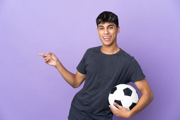 比赛一个年轻的足球运动员站在隔离的紫色墙壁上 用手指着旁边 展示一个产品休闲看球员