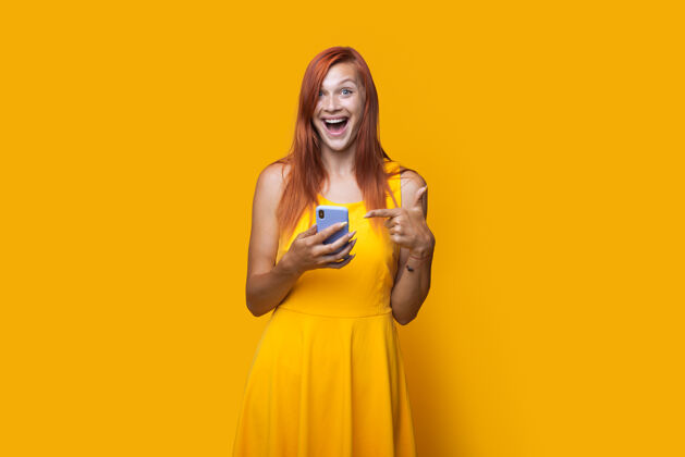 姜黄色工作室墙上 一位穿着裙子 红头发的白人妇女指着手机的单色照片技术生活方式电话