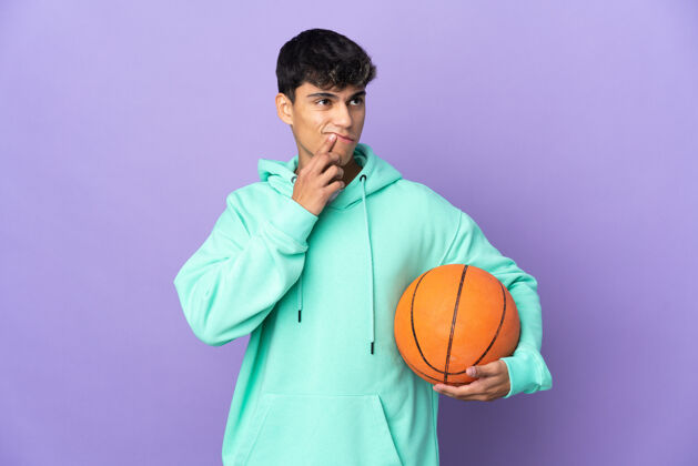 运动在孤立的紫色背景上打篮球的年轻人抬头时有疑惑制服沉思游戏