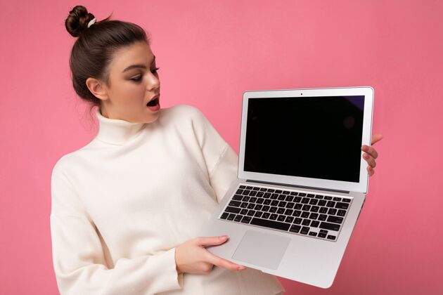 互联网照片中 一位美丽而惊讶的女士 一头浓密的黑发 穿着白色毛衣 手里拿着一台笔记本电脑 看着粉红色背景上的一本打开的上网本屏幕学生女孩