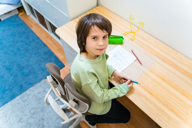 可爱一个年轻的男生坐在手术台的矫形椅上做作业家集中儿童写作练习很高兴在家上学概念男孩知识房间
